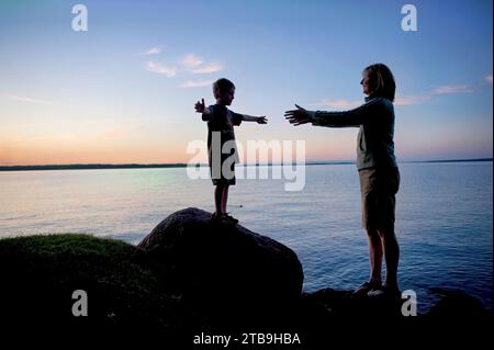 Mère aide son jeune fils à descendre d'un rocher le long du lac Leech au coucher du soleil ; Walker, Minnesota, États-Unis d'Amérique Banque D'Images