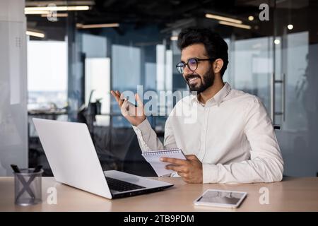 Souriant jeune homme d'affaires indien tenant la vidéoconférence, réunion de travail en ligne via appel vidéo sur ordinateur portable. Il parle, fait des gestes avec ses mains. Banque D'Images