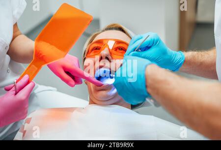 Jeune femme dans les lunettes de protection pendant la procédure de polymérisation de matériau d'obturation composite de dents avec la lumière UV de durcissement au cabinet du dentiste. Santé Banque D'Images
