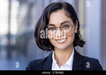 Photo en gros plan d'une jeune belle avocate réussie dans un costume et des lunettes souriant à la caméra et debout à l'extérieur. Banque D'Images