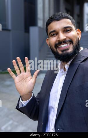 Photo en gros plan vertical d'un jeune homme d'affaires musulman souriant parlant au téléphone et agitant à la caméra, debout à l'extérieur d'un centre de bureaux. Banque D'Images