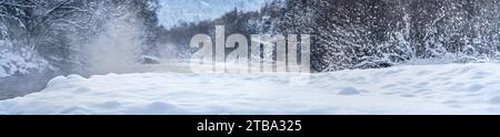 Rivière froide d'hiver, vapeur visible au-dessus de l'eau, arbres sombres des deux côtés, rivage couvert de neige fraîche au premier plan - large panorama Banque D'Images