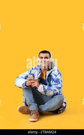 Jeune homme dans la veste chaude avec des écouteurs à l'aide d'un téléphone portable sur fond jaune Banque D'Images