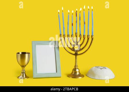 Menorah, kippah, cadre d'image vierge et tasse pour la célébration Hanukkah sur fond jaune Banque D'Images