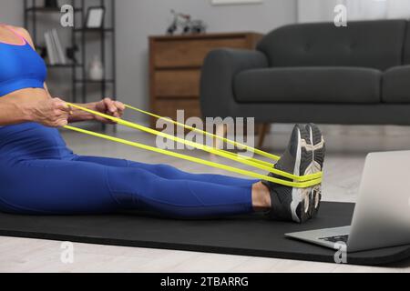 Femme faisant de l'exercice avec la bande élastique de fitness près de l'ordinateur portable sur le tapis à la maison, closeup Banque D'Images