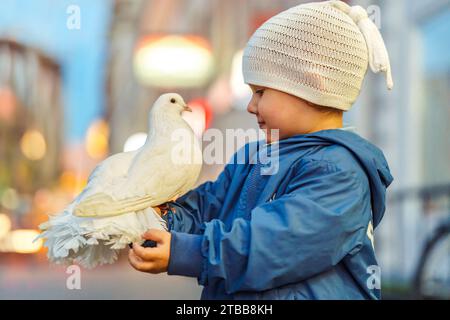 Heureux petit garçon avec pigeon blanc assis sur le bras en ville. Enfant en bas âge souriant avec pigeon entraîné sur la rue de la ville. Oiseau animal de compagnie sur la main d'enfant Banque D'Images