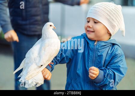 Heureux petit garçon avec pigeon blanc assis sur le bras en ville. Enfant en bas âge souriant avec pigeon entraîné sur la rue de la ville. Oiseau animal de compagnie sur la main d'enfant Banque D'Images
