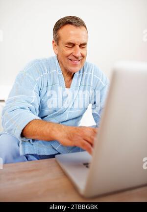 Maison, peignoir et homme âgé avec un ordinateur portable, dactylographie et Internet avec des informations de site Web, sourire et se détendre. Appartement, gars mature et personne avec un pc Banque D'Images