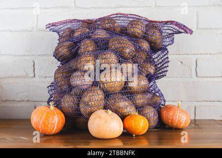 Au milieu du charme rustique d'une table en bois, un sac en maille déborde de pommes de terre fraîches et de citrouilles mûres, symbolisant la récolte abondante et abondante Banque D'Images