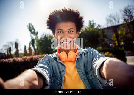 Portrait de beau mec joyeux avec chevelure porter des écouteurs de chemise en denim élégant blog faisant selfie souriant dans le parc à l'extérieur Banque D'Images