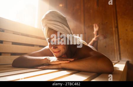 Femme se détendant dans un sauna finlandais, avec une serviette sur la tête, souriant à la caméra Banque D'Images