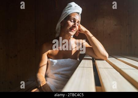 Femme dans un sauna finlandais penchée en arrière, les yeux fermés, avec une serviette enroulée autour de sa tête, éclairée par le soleil dans un centre thermal Banque D'Images