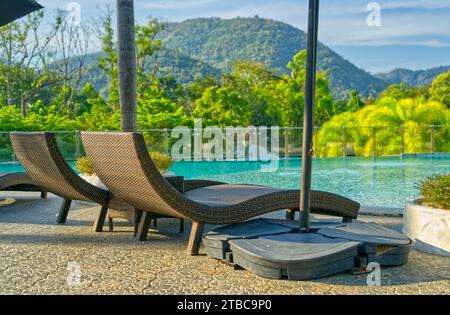Vider deux chaises longues en osier à côté d'une piscine d'une station, lumière du matin, fond d'arbres, montagne et ciel. Banque D'Images