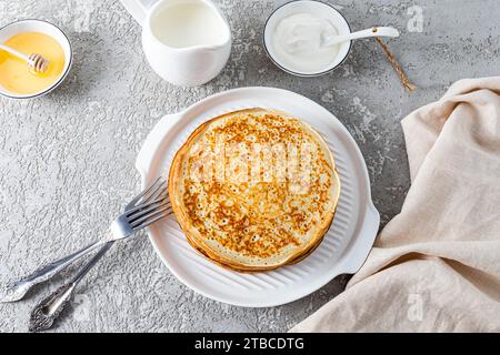 Vue de dessus d'une pile de délicieuses crêpes sur une assiette en relief blanc, couverts, crème sure, miel dans des bols. Petit déjeuner traditionnel festif. Banque D'Images