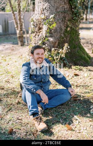 Jeune homme souriant assis sur la pelouse ensoleillée près de l'arbre Banque D'Images