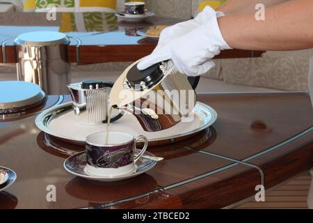 Image en gros plan d'un thé/café versé dans une tasse avec soucoupe à partir d'une théière en argent par une hôtesse de l'air portant des gants blancs. Banque D'Images