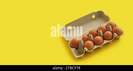 Œufs de poule bruns rustiques et biologiques dans un plateau en carton durable ouvert (boîte) sur un fond jaune monochrome lumineux. Image festive ludique Banque D'Images