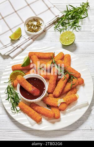 bâtonnets de mozzarella panés frits servis avec sauce, tranches de citron vert et romarin frais sur assiette blanche sur table en bois blanc avec ingrédients, verti Banque D'Images