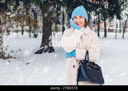 Heureuse femme souriante marchant dans le parc d'hiver enneigé portant manteau blanc de fourrure de peau de mouton tenant sac à main appréciant le paysage Banque D'Images