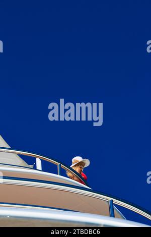 Fille portant une robe rouge et un chapeau de paille debout sur le pont latéral d'un superyacht tout en tenant sur le rail en acier inoxydable avec un ciel bleu profond. Banque D'Images