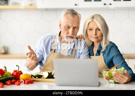 Mauvaise recette. Conjoints aînés confus regardant un ordinateur portable tout en cuisinant dans la cuisine Banque D'Images