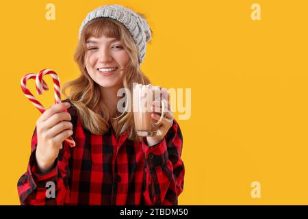 Jolie jeune femme tenant une tasse de chocolat chaud avec des guimauves et des cannes de bonbons sur fond jaune Banque D'Images