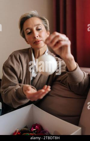 Portrait vertical d'une femme d'âge moyen tenant et regardant boule de Noël festive avec une expression heureuse. La femelle élégante décore l'arbre de Noël pour New Banque D'Images