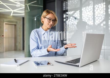 La travailleuse en lunettes est assise à une table devant un ordinateur portable, communique par appel vidéo, conseille les clients, fait beaucoup de gestes. Banque D'Images