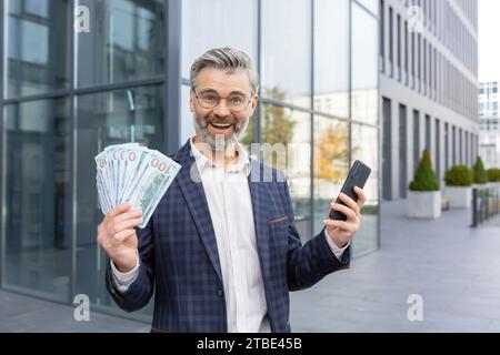 Homme d'affaires surpris tenant pile d'argent et téléphone dans les mains, heureux avec la loterie gagnante, la promotion au travail, regardant la caméra et souriant. Banque D'Images