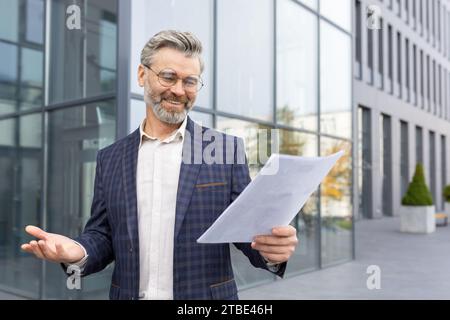 Homme d'affaires satisfait en costume tenant des documents d'affaires, debout devant le bureau dans la rue, négociation réussie, bonne nouvelle. Banque D'Images