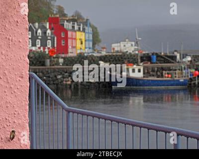 Mur rose et balustrade en métal avec des bâtiments colorés côté port au-delà de la jetée avec des pots de crabe et bateau de pêche sous une pluie légère - Tobermory, Mull, Écosse, Royaume-Uni Banque D'Images