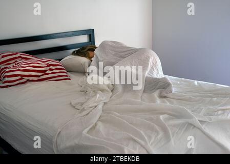 Femme dormant sous des draps de lin blanc à la lumière du jour avec des oreillers striés rouges et blancs sur la gauche Banque D'Images