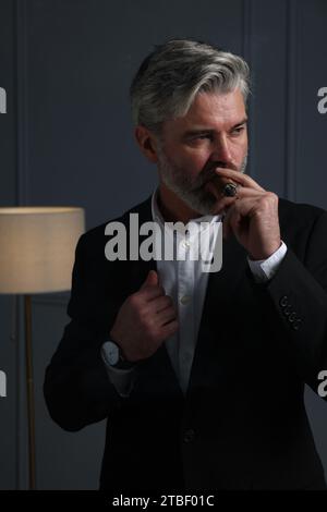 Bel homme barbu fumant le cigare près du mur gris foncé à l'intérieur Banque D'Images