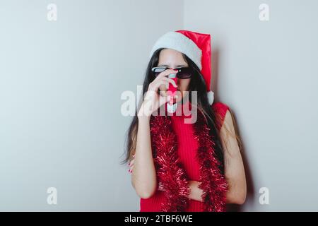 Fille cool-asthmatique à Noël. Jeune femme avec asthme portant le chapeau du Père Noël, décorations de Noël et lunettes de soleil tout en utilisant son inhalateur Banque D'Images