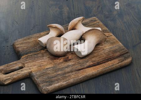 Champignons eryngi frais, varient en tailles, reposent sur une planche à découper en bois, fond gris-bleu surface en bois teinté, scène vibrante de la préparation des aliments Banque D'Images