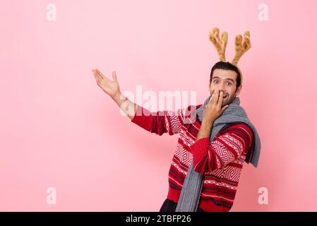 Le studio de Noël a photographié le portrait d'un homme caucasien choqué portant un chandail et un bandeau de renne de fantaisie pointant la main vers le haut pour copier l'espace en isolat de couleur rose Banque D'Images