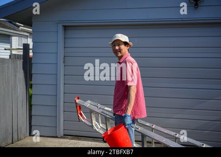 Homme tenant une échelle et un seau rouge après avoir fini le travail. Travaux d'entretien de la maison. Auckland. Banque D'Images