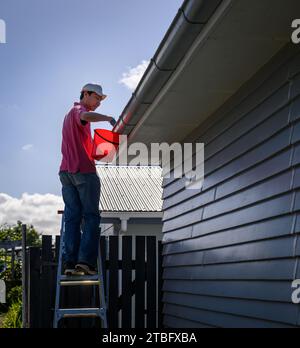 Homme debout sur l'échelle et nettoyant la gouttière. Travaux d'entretien de la maison. Format vertical. Auckland. Banque D'Images