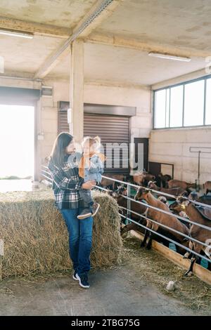 Mère avec une petite fille dans les bras se tient appuyée sur une botte de foin près d'un paddock de chèvres Banque D'Images