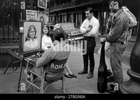 Junge Männer sehen einem Porträtzeichner im French Quarter dabei zu, wie er das Bildnis einer jungen Frau mit Pastellkreiden anfertigt, Nouvelle-Orléans 1965. De jeunes hommes regardent un portraitiste dans le quartier français faire un portrait d'une jeune femme au pastel, Nouvelle-Orléans 1965. Banque D'Images