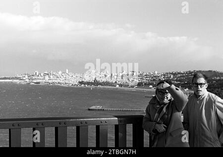 Ein Mann und eine Frau auf der Golden Gate Bridge, im hintergrund die Torpedo Wharf sowie die Skyline von San Francisco, 1962. Un homme et une femme sur le Golden Gate Bridge, avec Torpedo Wharf et la Skyline de San Francisco en arrière-plan, 1962. Banque D'Images