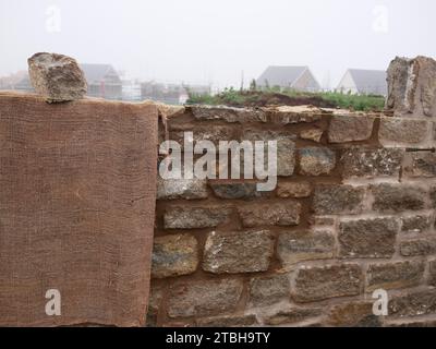 Mur d'enceinte avec façade traditionnelle en pierre en cours de construction dans un nouveau développement immobilier. Hessian protège la nouvelle maçonnerie du froid. Banque D'Images