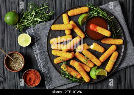 bâtonnets de mozzarella panés et frits servis avec sauce, tranches de citron vert et romarin frais sur plateau noir sur table en bois noir, cuisine américaine Banque D'Images