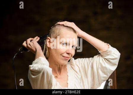 Une femme sourit et se coupe les cheveux sur la tête avec une tondeuse sur un fond sombre. Banque D'Images