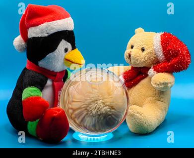 Pingouin bleu foncé et ours en peluche beige , assis ensemble prédisant l'avenir sur boule de cristal. Animaux en peluche isolés sur fond de papier bleu. Banque D'Images