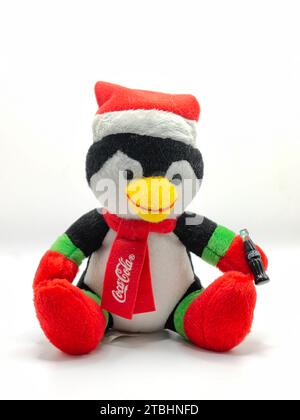 Pingouin bleu foncé isolé, sur fond de papier blanc. Porter un chapeau de noël rouge et une écharpe avec logo coca cola dessus. Peluche jouet animal.Joyeuses fêtes. Banque D'Images