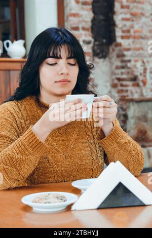 image verticale d'une jeune femme latine assise buvant du café avec prudence car il fait très chaud, tenant une tasse en porcelaine avec ses mains. Banque D'Images