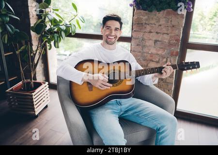 Photo de positif funky jeune gars habillé chemise blanche jouant de l'instrument acoustique à l'intérieur de la salle de la maison Banque D'Images