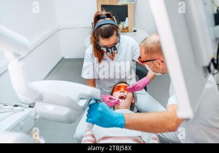 Rendez-vous en clinique dentaire. Femme dentiste en loupe faisant la prévention des dents à l'aide d'outils médicaux. Jeune homme assistant pointant la lumière. Banque D'Images