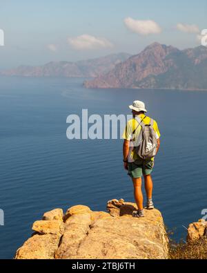 Marcheur au sommet d'un rocher observant la mer au sentier de la Tête de chien, Piana, Corse, France. Banque D'Images